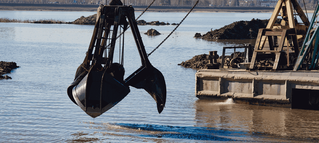Clamshell dredging bucket lifting sediment from a river beside a barge during hydraulic dredging operations, supporting waterway maintenance, sediment removal, channel deepening, shoreline stabilization, and environmental restoration for marine construction and coastal infrastructure projects.