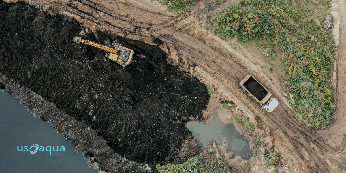 A yellow excavator and a dump truck at an industrial construction site near a body of water using a marsh buggy provided by U.S. Aqua