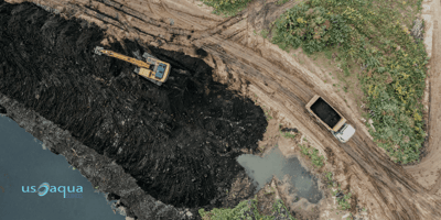 A yellow excavator and a dump truck at an industrial construction site near a body of water using a marsh buggy provided by U.S. Aqua