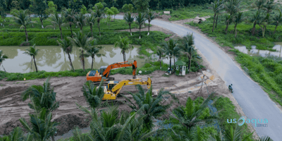 Two excavators, one yellow and one orange, in a tropical environment  next to a road 